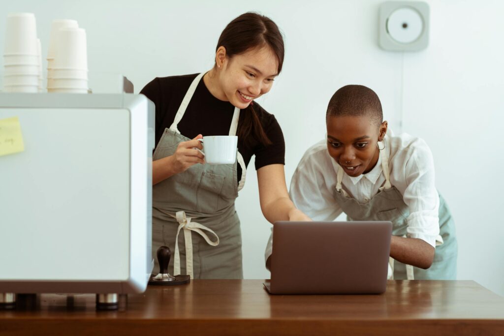 Joyful smiling multiracial waitresses surfing internet on contemporary netbook and smiling while having coffee break in cozy kitchen