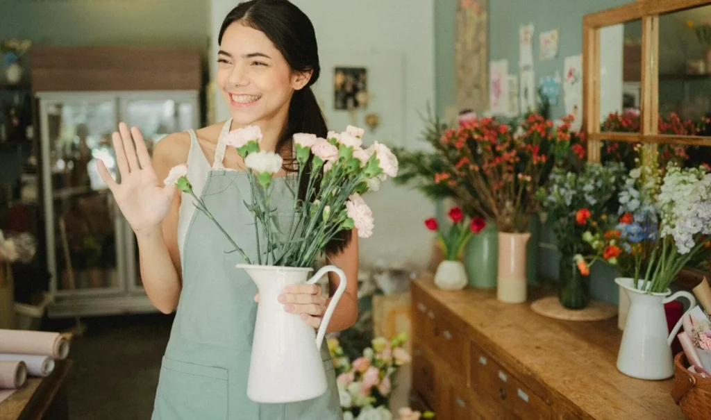 Friendly young florist waving in her shop, holding a bouquet of flowers.